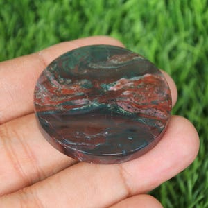 May include: A round, polished gemstone with a mix of colors. The stone features a dark green base with swirling patterns of red and brown. The gemstone is held in a hand against a backdrop of green grass.