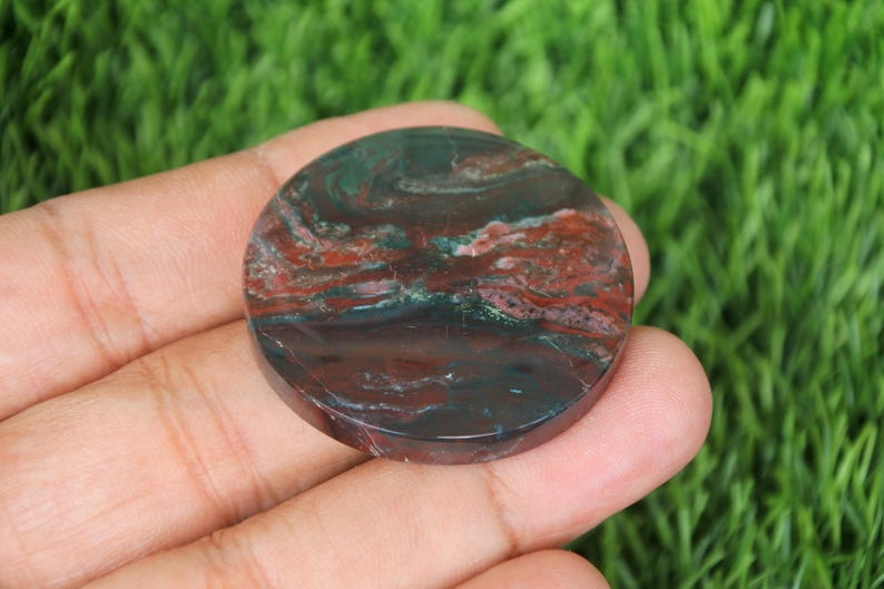 May include: A round, polished gemstone with a mix of colors. The stone features a dark green base with swirling patterns of red and brown. The gemstone is held in a hand against a backdrop of green grass.