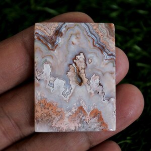 May include: A rectangular polished gemstone cabochon featuring intricate, swirling patterns in shades of cream, white, soft pink, and earthy brown. The stone displays a natural, banded agate structure with translucent and opaque areas throughout.