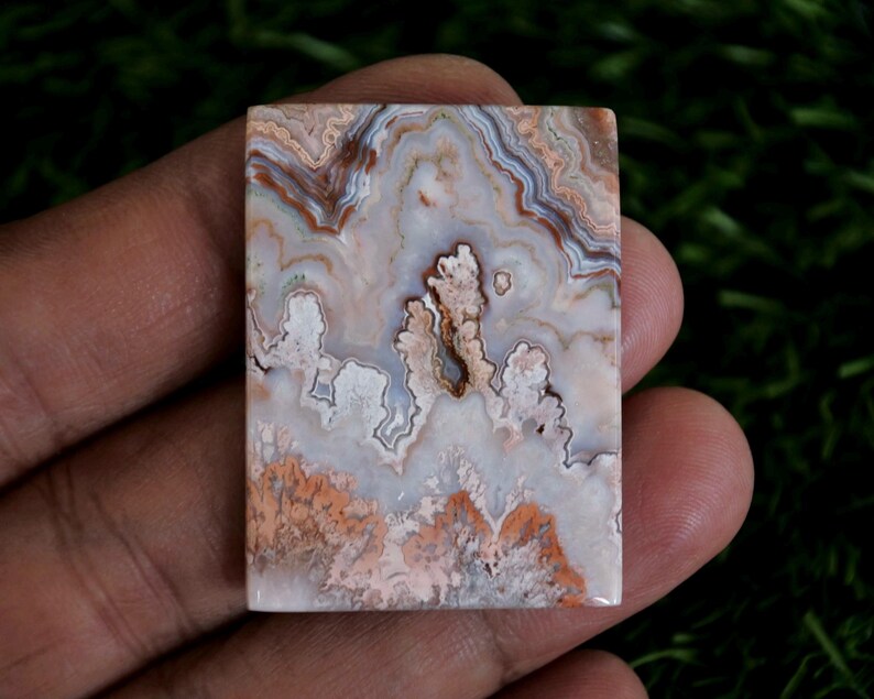 May include: A rectangular polished gemstone cabochon featuring intricate, swirling patterns in shades of cream, white, soft pink, and earthy brown. The stone displays a natural, banded agate structure with translucent and opaque areas throughout.