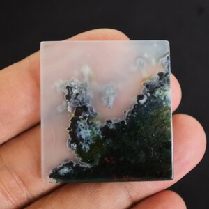 Puede incluir: Gema cuadrada con un fondo rosa p&aacute;lido y un patr&oacute;n similar a un paisaje de color verde oscuro, negro y blanco. La piedra se sostiene en una mano, mostrando su superficie pulida y dise&ntilde;o natural.