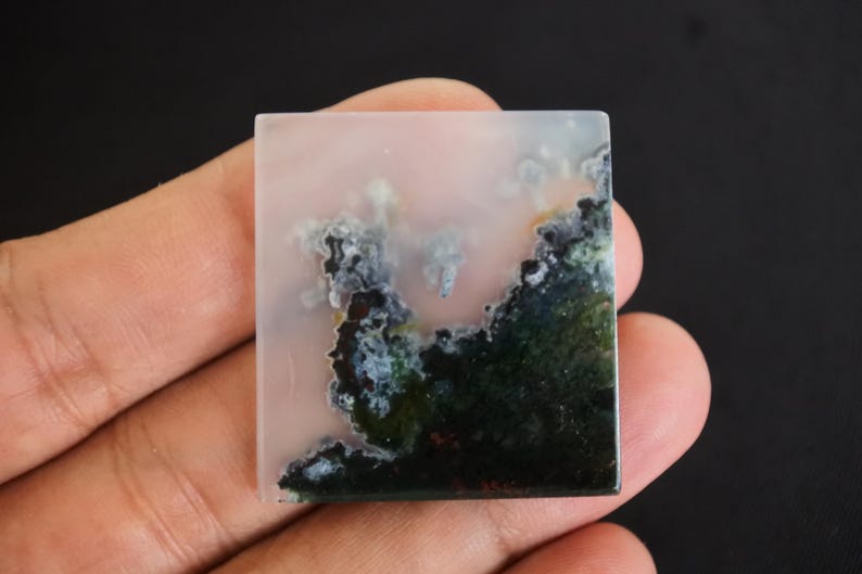 Puede incluir: Gema cuadrada con un fondo rosa p&aacute;lido y un patr&oacute;n similar a un paisaje de color verde oscuro, negro y blanco. La piedra se sostiene en una mano, mostrando su superficie pulida y dise&ntilde;o natural.