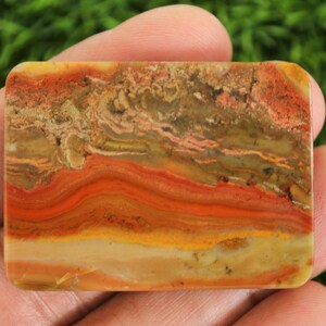 Puede incluir: Una piedra preciosa rectangular con una superficie pulida. La piedra presenta capas de tonos naranja, rojo, amarillo y marr&oacute;n, creando un patr&oacute;n natural y terroso. Los bordes son redondeados y la apariencia general es suave y t&aacute;ctil.