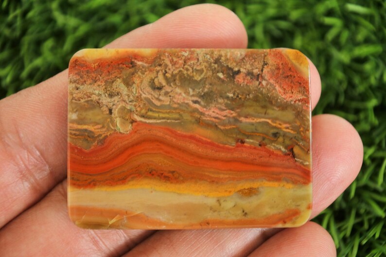 Puede incluir: Una piedra preciosa rectangular con una superficie pulida. La piedra presenta capas de tonos naranja, rojo, amarillo y marr&oacute;n, creando un patr&oacute;n natural y terroso. Los bordes son redondeados y la apariencia general es suave y t&aacute;ctil.