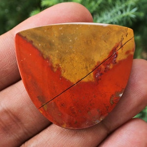 K&ouml;nnte beinhalten: Ein roter, gelber und brauner Jaspis-Cabochon mit einem nat&uuml;rlichen Riss, der durch ihn verl&auml;uft. Der Cabochon hat eine Tropfenform und eine polierte Oberfl&auml;che.