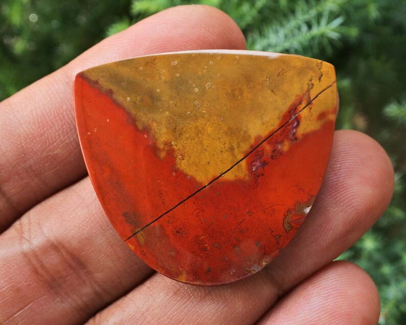 K&ouml;nnte beinhalten: Ein roter, gelber und brauner Jaspis-Cabochon mit einem nat&uuml;rlichen Riss, der durch ihn verl&auml;uft. Der Cabochon hat eine Tropfenform und eine polierte Oberfl&auml;che.