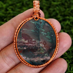 May include: A handcrafted pendant featuring a polished, round stone encased in copper wire. The stone displays a mix of green, maroon, and grey tones. The copper wire is intricately woven, creating a unique piece of jewellery.