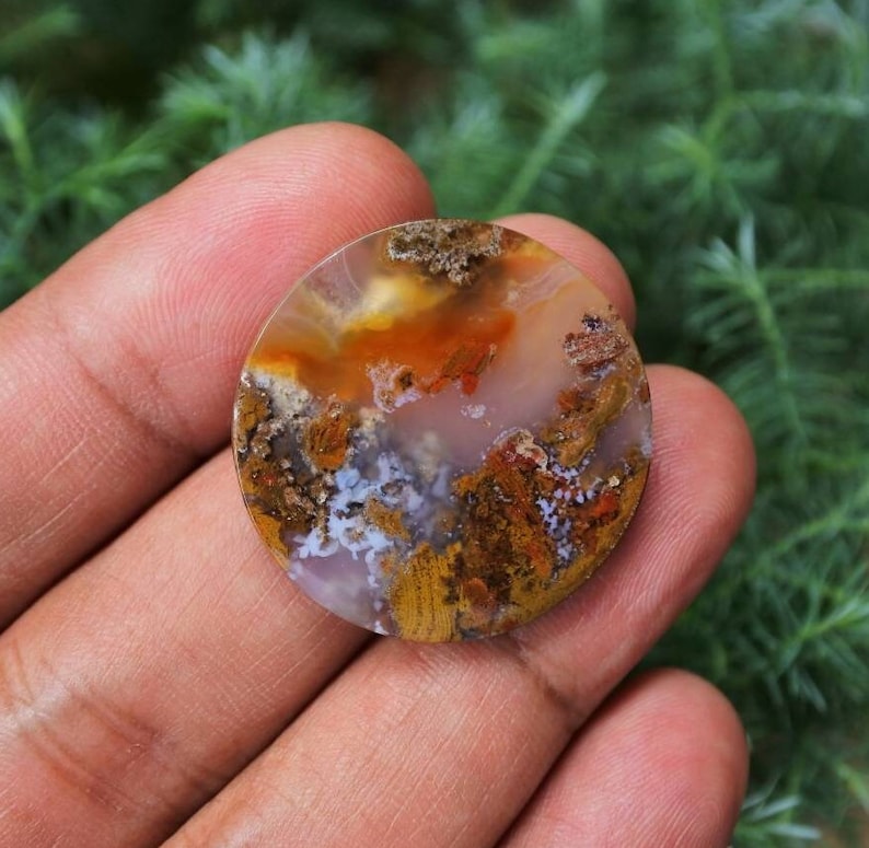 May include: A round, polished agate cabochon featuring a range of colours, including white, brown, orange, and red. The agate displays a distinctive, swirling pattern.