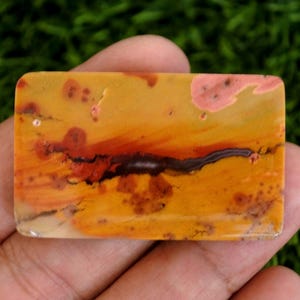 May include: A rectangular polished gemstone cabochon featuring a warm color palette of golden yellow, burnt orange, and deep red tones. The stone displays natural organic patterns with dark brown linear inclusions and soft pinkish spots.