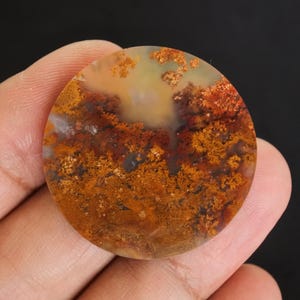 Puede incluir: Una gema redonda y pulida con un patr&oacute;n natural y terroso. La piedra presenta una mezcla de colores c&aacute;lidos, incluyendo tonos de naranja, marr&oacute;n y amarillo, creando un dise&ntilde;o &uacute;nico y org&aacute;nico. La gema se sostiene en una mano.