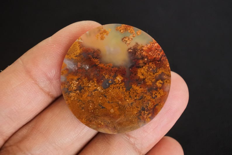 Puede incluir: Una gema redonda y pulida con un patr&oacute;n natural y terroso. La piedra presenta una mezcla de colores c&aacute;lidos, incluyendo tonos de naranja, marr&oacute;n y amarillo, creando un dise&ntilde;o &uacute;nico y org&aacute;nico. La gema se sostiene en una mano.