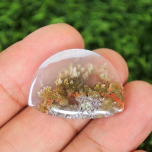 Pode incluir: Uma pedra preciosa translúcida, lapidada em cabochão, com um padrão semelhante a uma paisagem. A pedra apresenta uma base clara com inclusões castanhas, laranja e cinza, assemelhando-se a uma floresta ou paisagem em miniatura. A pedra é segurada na mão.