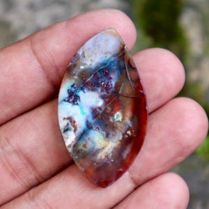 May include: A polished, teardrop-shaped gemstone with a unique pattern of colors. The stone features a mix of white, blue, red, and brown hues, with intricate veining. The gemstone is held in a hand, showcasing its natural beauty.