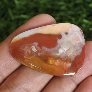 May include: A polished, teardrop-shaped cabochon stone featuring a swirling pattern of earthy colors including deep terracotta red, creamy beige, translucent white, and golden yellow. The surface is smooth and glossy, reflecting light across its varied mineral inclusions and organic, marbled color transitions.
