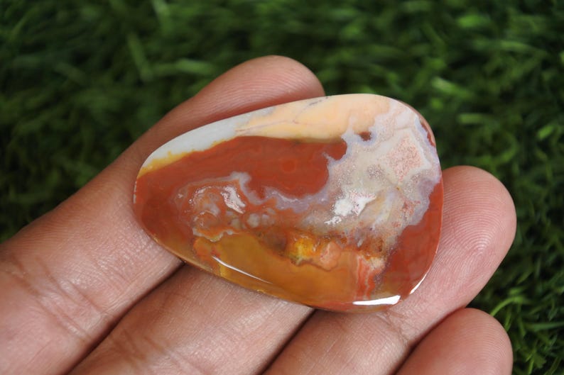 May include: A polished, teardrop-shaped cabochon stone featuring a swirling pattern of earthy colors including deep terracotta red, creamy beige, translucent white, and golden yellow. The surface is smooth and glossy, reflecting light across its varied mineral inclusions and organic, marbled color transitions.
