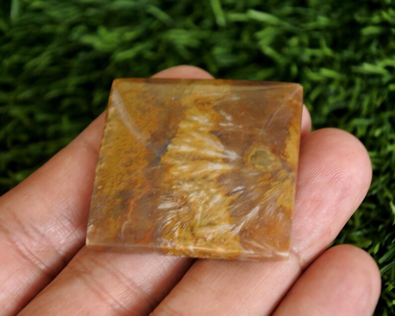 May include: A square-shaped polished stone cabochon featuring a warm colour palette of golden brown, tan, and cream. The surface displays intricate, feathery, and fibrous white inclusions that create a natural, swirling pattern across the flat face.