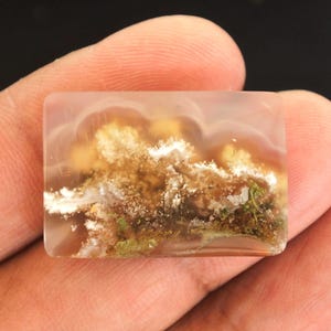 Peut inclure: Une pierre précieuse rectangulaire et translucide avec un motif de paysage. La pierre présente un mélange de teintes marron, blanches et vertes, ressemblant à des nuages et à de la végétation. La pierre est tenue entre deux doigts.