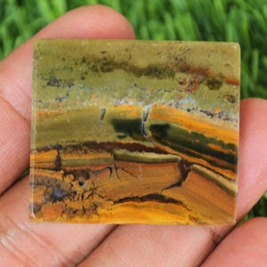 May include: Square gemstone with a natural, earthy appearance. The stone features layers of olive green, golden yellow, and brown hues, with intricate patterns and textures. The gemstone is held in a hand.