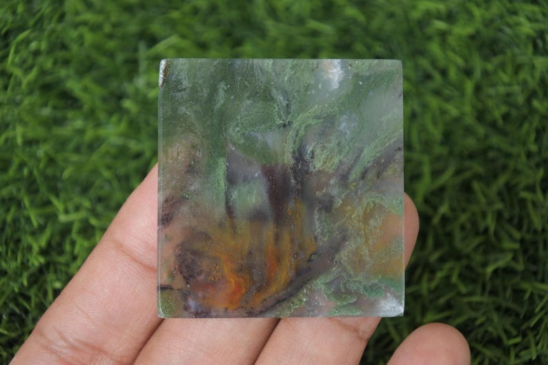 Peut inclure: Une pierre pr&eacute;cieuse carr&eacute;e et polie, pr&eacute;sentant un motif unique de teintes vertes, orange et brunes. La surface de la pierre a un aspect marbr&eacute;, avec des couleurs et des textures tourbillonnantes. La pierre est tenue dans une main sur un fond vert.