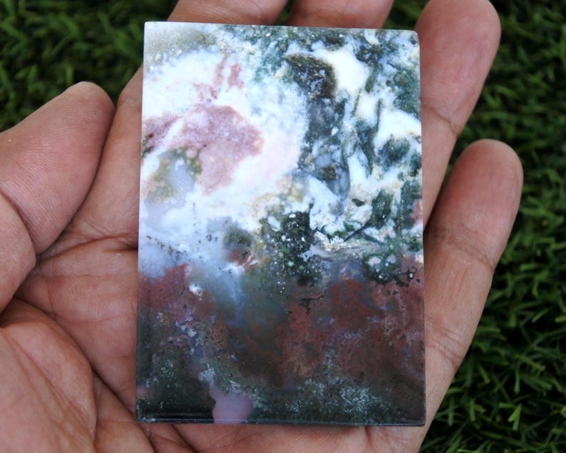 K&ouml;nnte beinhalten: Ein rechteckiger Stein mit einem einzigartigen Farbmuster. Der Stein weist eine Mischung aus wei&szlig;en, rosa, gr&uuml;nen und braunen Farbt&ouml;nen auf. Der Stein wird in einer Hand gehalten, mit einem gr&uuml;nen Hintergrund.