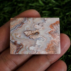 May include: A rectangular polished stone cabochon featuring intricate banding and swirling patterns in shades of cream, white, soft pink, and earthy brown. The surface displays a smooth, glossy finish with distinct mineral layers and organic textures.
