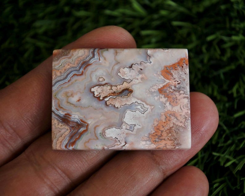 May include: A rectangular polished stone cabochon featuring intricate banding and swirling patterns in shades of cream, white, soft pink, and earthy brown. The surface displays a smooth, glossy finish with distinct mineral layers and organic textures.