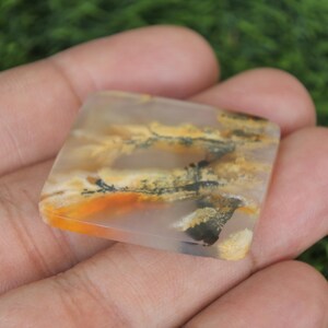 May include: A square-shaped, translucent gemstone with a landscape-like pattern of orange, yellow, and black inclusions. The stone is held in a hand against a blurred green background, showcasing its natural beauty and unique design.