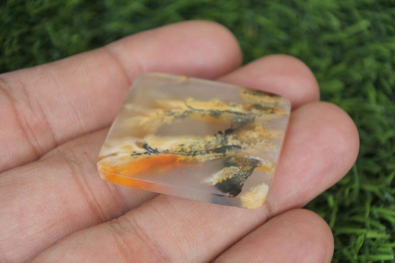 May include: A square-shaped, translucent gemstone with a landscape-like pattern of orange, yellow, and black inclusions. The stone is held in a hand against a blurred green background, showcasing its natural beauty and unique design.