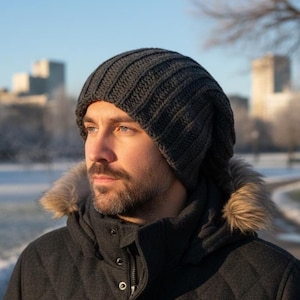May include: A dark grey, ribbed knit beanie hat. The hat is worn with a dark grey quilted jacket with a fur-lined hood. The background shows a city skyline on a bright, sunny day.