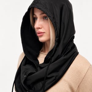 May include: A black hooded neck warmer featuring a smooth, water-resistant outer shell and a soft interior lining. The hood includes a drawstring for adjustment and a front placket secured with two black snap buttons at the base of the neck.