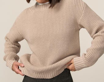 May include: A beige, ribbed knit sweater with a mock neck and long sleeves. The sweater is paired with dark gray, high-waisted pants. The model is wearing a white undershirt. The sweater has a relaxed fit.