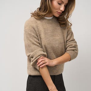 May include: A tan-colored, ribbed knit sweater with long sleeves. The sweater is paired with dark gray pants. The model is wearing gold rings. The sweater has a crew neck and is made of a soft, textured material.