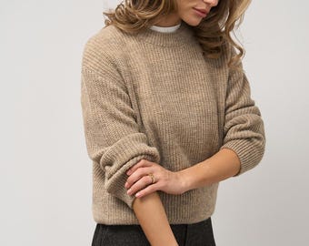 May include: A tan-colored, ribbed knit sweater with long sleeves. The sweater is paired with dark gray pants. The model is wearing gold rings. The sweater has a crew neck and is made of a soft, textured material.
