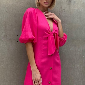 May include: A bright pink midi dress with a tie front and a button-up closure. The dress has puffed sleeves and a v-neckline.