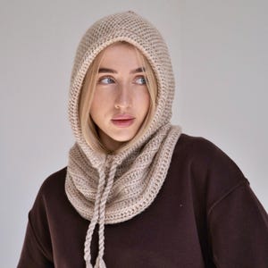 May include: A beige knitted balaclava hood featuring a textured ribbed stitch pattern and a thick braided drawstring cord at the neck. The head covering is crafted from a neutral-toned yarn and provides a snug fit around the face and neck area.