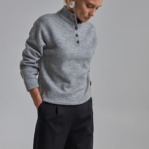 Op de afbeelding: Een grijze trui met lange mouwen, een hoge kraag en drie zwarte knopen. Het model draagt een zwarte broek. De trui is gemaakt van een zacht, getextureerd materiaal. De outfit is eenvoudig en elegant.