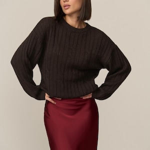 May include: A woman wearing a dark brown, ribbed knit sweater and a burgundy satin skirt. The sweater has a crew neck and long sleeves. The skirt is a solid color and has a slight sheen. The outfit is a mix of textures and colors.