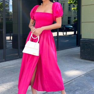 May include: A woman wearing a bright pink, sleeveless dress with a square neckline and a side slit. She is carrying a white handbag with a gold handle and wearing white heeled sandals.