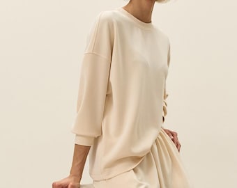 May include: A cream-colored long-sleeve sweatshirt with a crew neckline and matching cream-colored trousers. The sweatshirt features dropped shoulders and ribbed cuffs, while the trousers have a relaxed fit with a gathered, elasticized waistband.