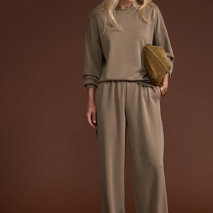May include: An olive green lounge set comprising a crew neck sweatshirt and wide-leg trousers. The outfit is accessorised with a matching clutch. The model is wearing sunglasses.