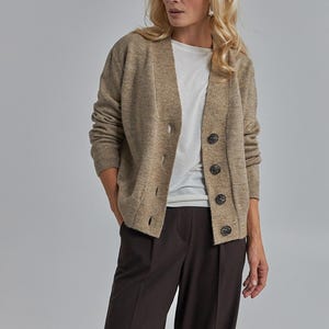 May include: A woman wearing a beige cardigan with large dark buttons over a white t-shirt and dark brown wide-leg pants. The cardigan has a relaxed fit. The background is a solid light gray.