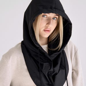 May include: A black hooded neck warmer featuring a quilted texture, a rounded hemline, and a front placket with snap button closures. The hood includes integrated drawstrings with aglets, providing a structured fit around the head and neck area.