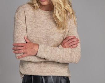 May include: A long-sleeved, crew-neck sweater in a heathered beige knit fabric, paired with a black skirt featuring a smooth, semi-glossy finish. The sweater has a relaxed fit with ribbed cuffs and a ribbed neckline, while the skirt is pleated.