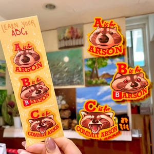 Puede incluir: Un conjunto de pegatinas y un marcapáginas con temática de mapache. El marcapáginas dice "Learn Your ABCs" con "A is for ARSON", "B is for ARSON" y "C is for COMMIT ARSON". Pegatinas adicionales muestran el mapache con las letras A, B y C.