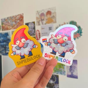 May include: Two colourful gnome stickers. One is yellow, the other is holographic. Both feature a cartoon gnome with a red hat, white beard, and the text "SHMEBULOCK". The holographic sticker has a rainbow flag.