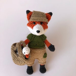 May include: A crocheted fox wearing a brown hat and green shirt, holding a brown basket with a white crocheted goose inside.