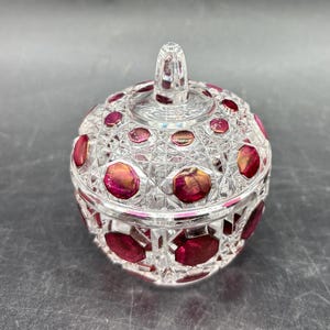 May include: A clear, round crystal jar with a lid, featuring ruby red accents. The jar has a cut-glass design with red circular inserts. The lid has a small, clear handle. The jar is likely used for storing small items.