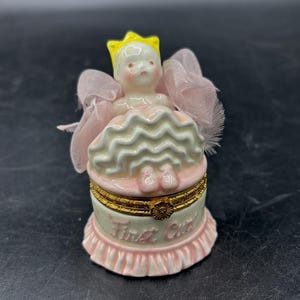 May include: A small, decorative trinket box with a porcelain doll figurine on top. The doll wears a yellow crown and a pink dress. The box is pink and white with gold accents and the words "First Child" in pink.