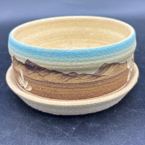 May include: A round ceramic planter with a matching saucer, featuring a textured beige base, a brown landscape design depicting mountains and cacti, and a bright blue rim. The interior is a smooth, light beige finish with a wide opening.