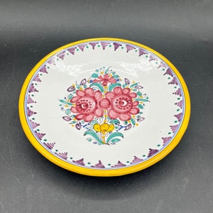 May include: A white ceramic plate with a yellow rim and a purple and green floral design. The flowers are pink and have a yellow center.